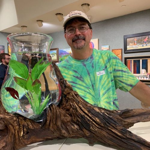 A man in a tie-dye shirt stands beside a decorative fish tank on a wooden base, with art in the background.