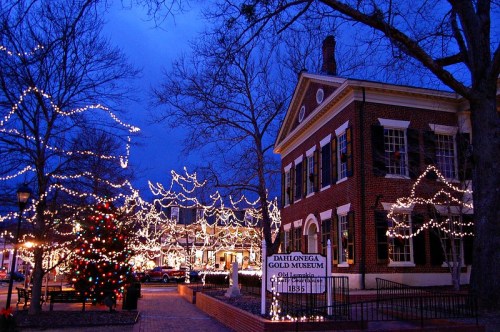 A historic building adorned with festive lights, surrounded by trees and a Christmas tree, set against a twilight sky.