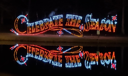 Colorful lights spell "Celebrate the Season," reflecting in a calm body of water at night.