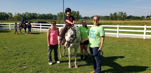 A child on a spotted horse, surrounded by three adults in a grassy area with a white fence and trees in the background.