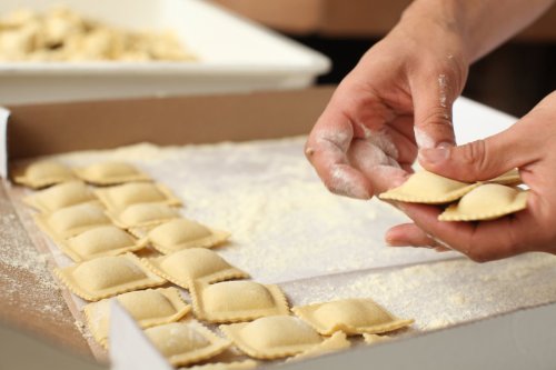 A person’s hands hold a ravioli while arranging uncooked ravioli on a floured surface in a box.