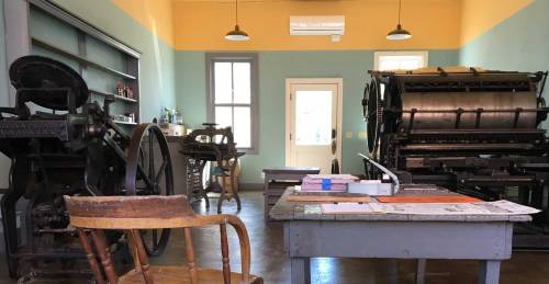 A vintage printing workshop with old printing presses, a wooden chair, and a table with papers and tools.