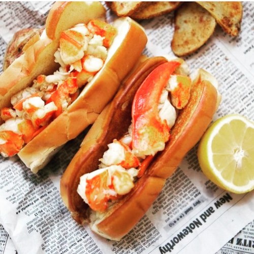 Two lobster rolls filled with lobster meat on toasted buns, served with lemon wedges and accompanied by potato wedges.