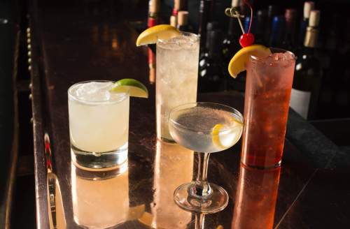Four colorful cocktails on a bar top, featuring garnishes of lemon and lime, with bottles of wine in the background.