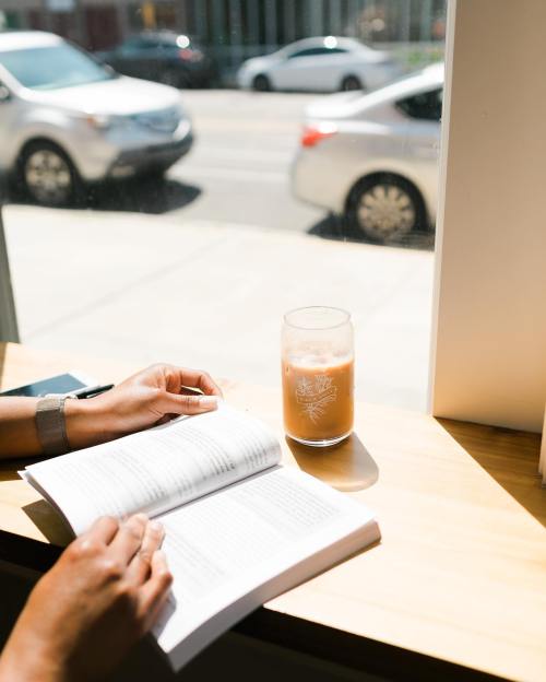 A person reading a book by a window with a glass of iced coffee, cars visible outside.