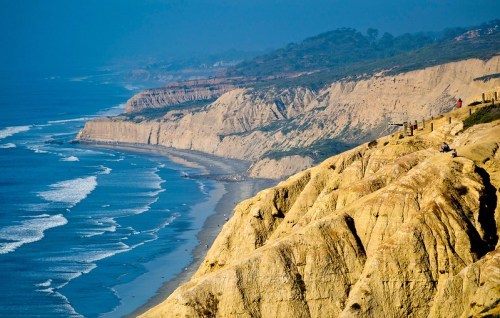 Coastal cliffs overlooking a sandy beach and ocean waves under a blue sky.