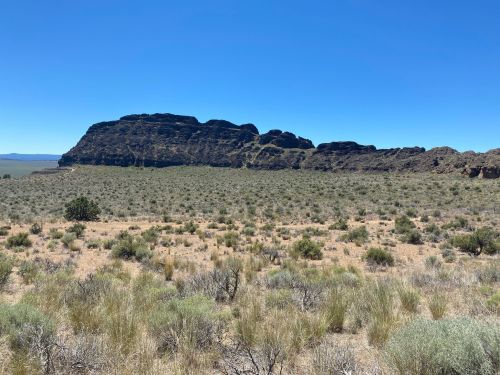 A rocky formation rises above a vast, arid landscape with sparse vegetation under a clear blue sky.