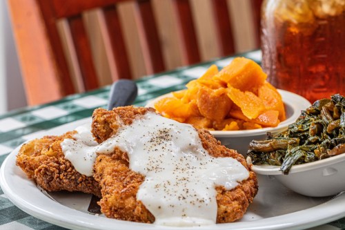 A plate of fried chicken with white gravy, collard greens, and sweet potatoes, served on a checkered tablecloth.