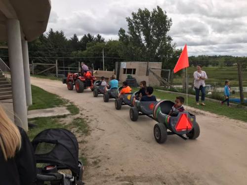 Children ride in small, colorful tractor carts along a dirt path, with adults supervising nearby.