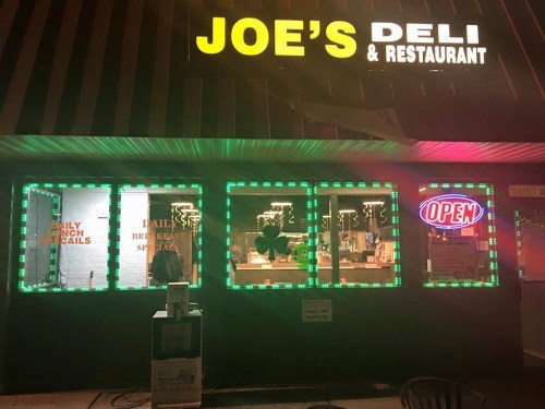 A brightly lit deli with neon signs reading "OPEN" and "DAILY LUNCH SPECIALS" in the windows at night.