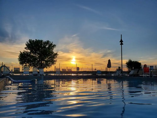 A serene pool at sunset, reflecting vibrant colors in the water, with lounge chairs and a tree in the background.