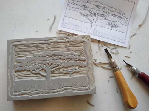 A carved block of clay depicting a tree, with tools and a sketch of the design nearby.