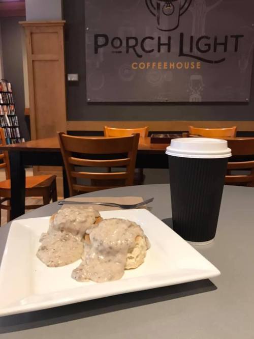 A plate of biscuits smothered in gravy next to a coffee cup in a cozy coffeehouse setting.