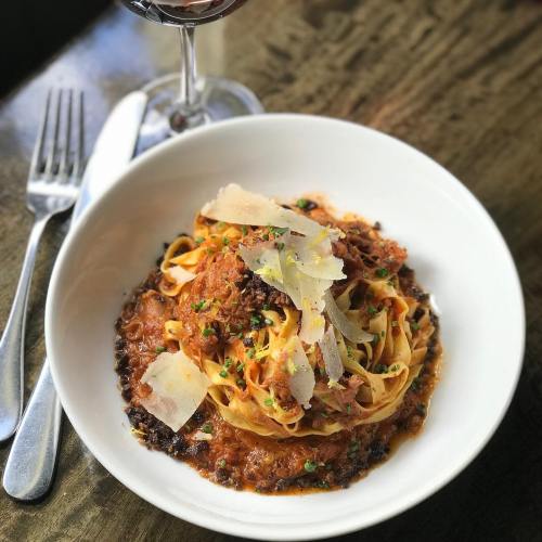 A bowl of pasta with rich sauce, topped with shredded meat and cheese, accompanied by a glass of red wine.