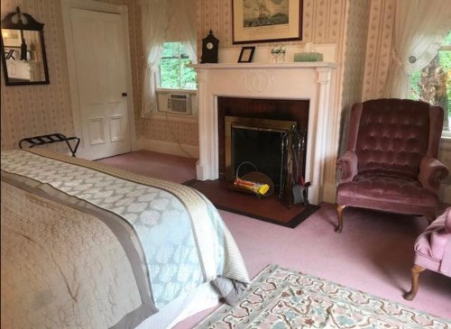Cozy bedroom with a bed, fireplace, armchair, and soft lighting, featuring patterned wallpaper and a warm atmosphere.