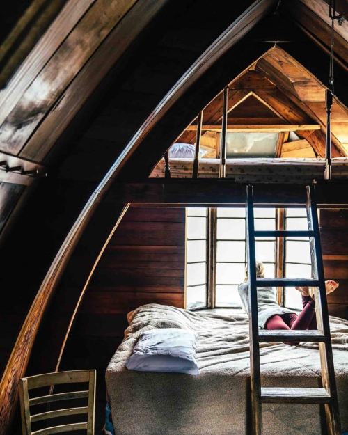 Cozy attic bedroom with wooden beams, a ladder, and a person sitting on the bed by a window.