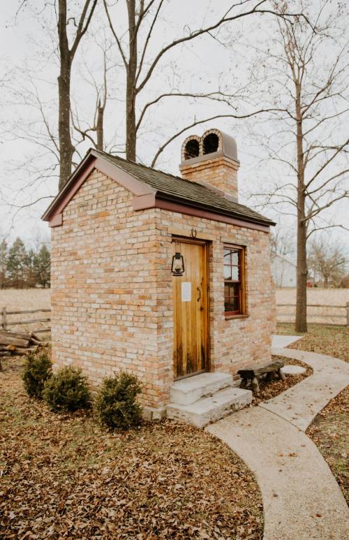 A small brick building with a chimney, surrounded by trees and a winding path, set in a rural landscape.