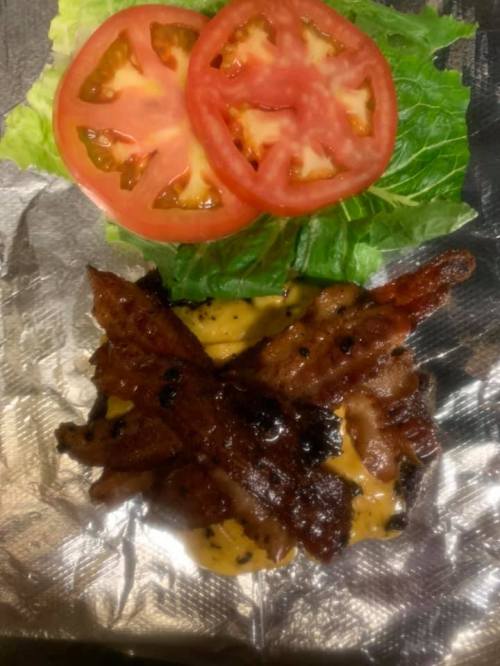 A serving of crispy bacon on a bed of melted cheese, topped with lettuce and tomato slices, wrapped in foil.