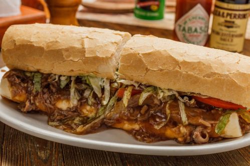 A hearty sandwich filled with beef, lettuce, and tomato, served on a crusty roll with sauces in the background.