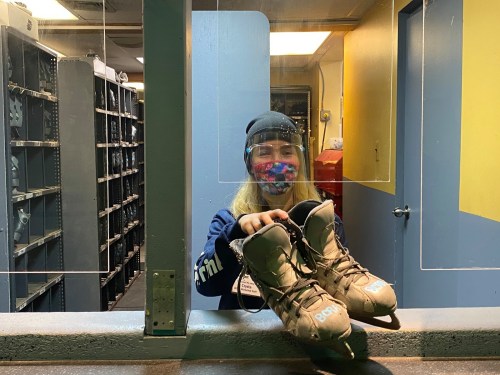 A person wearing a mask and beanie holds up a pair of shoes in a brightly lit room with shelves in the background.