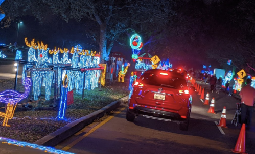A festive drive-through light display with colorful decorations, including reindeer and flamingos, along a lined road.