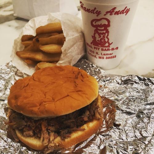 A pulled pork sandwich on a bun, served with fries and a drink from Handy Andy BBQ, all on a marble surface.