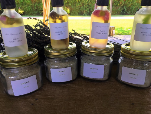 Bottles of essential oils and jars of detox salts displayed on a table, surrounded by greenery.