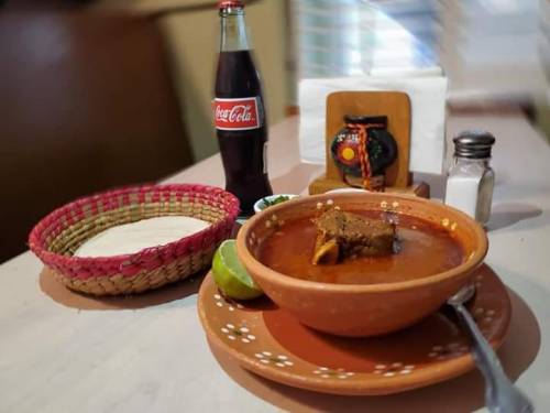 A bowl of soup with meat, lime, a basket of tortillas, a Coca-Cola bottle, and a salt shaker on a table.