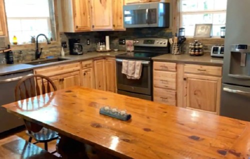 A rustic kitchen with wooden cabinets, a large table, and modern appliances, featuring natural light from windows.