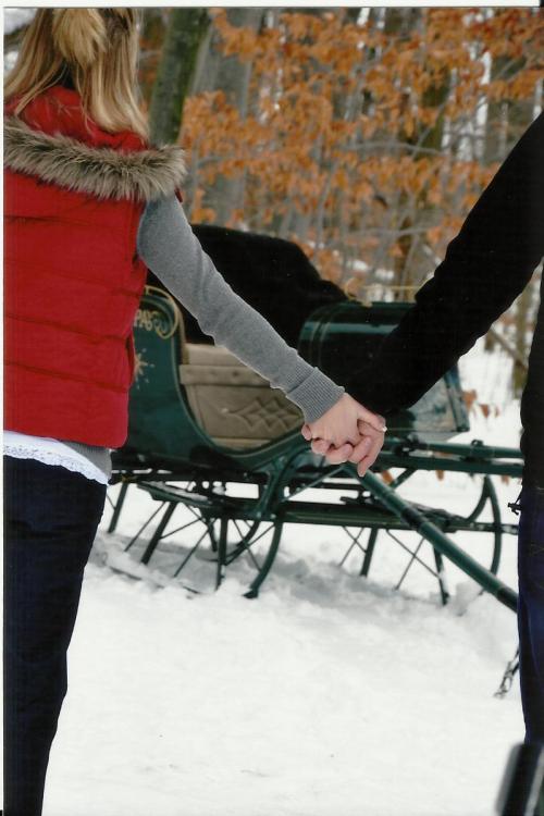 A couple holding hands in winter attire, with a vintage sleigh in a snowy forest background.