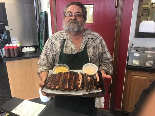 A smiling man in an apron holds a tray of ribs with two sides in a restaurant setting.