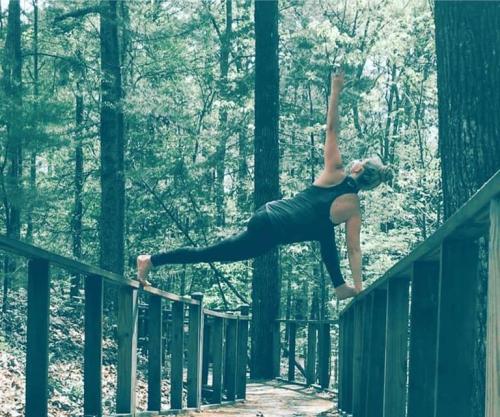 A person performs a yoga pose on a wooden railing in a forested area, surrounded by tall trees.