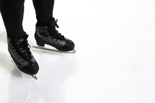 Close-up of a figure skater's feet in black skates on an ice rink, with blades glinting on the smooth ice surface.