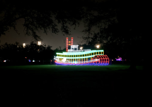 A brightly lit, colorful steamboat display in a dark park, surrounded by trees and distant lights.