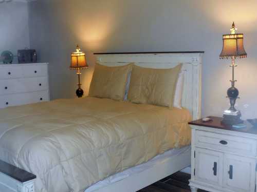 Cozy bedroom featuring a white bed with beige bedding, two lamps, and a dresser against a gray wall.