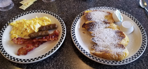 A plate with scrambled eggs, bacon, sausage links, and French toast dusted with powdered sugar and syrup.