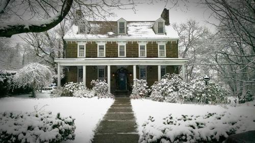 A large, historic house covered in snow, surrounded by trees and shrubs, with a clear path leading to the front door.