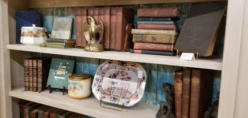 A wooden shelf displaying vintage books, decorative plates, and various antique items against a blue wooden background.