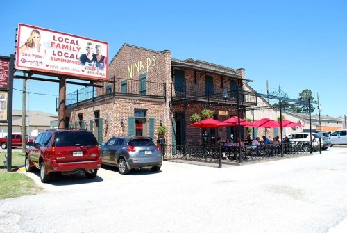 A brick building with outdoor seating and red umbrellas, featuring a sign for Nina's and a billboard for local businesses.