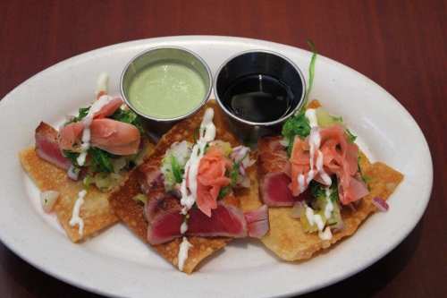 Three crispy wonton tacos topped with seared tuna, pickled ginger, and drizzled with sauces, served on a white plate.