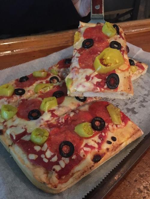 A tray of rectangular pizza topped with pepperoni, black olives, and green peppers, with a slice being lifted.