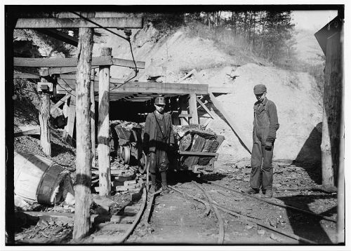 Two miners stand near a coal cart in a dimly lit mine, surrounded by wooden supports and rocky terrain.