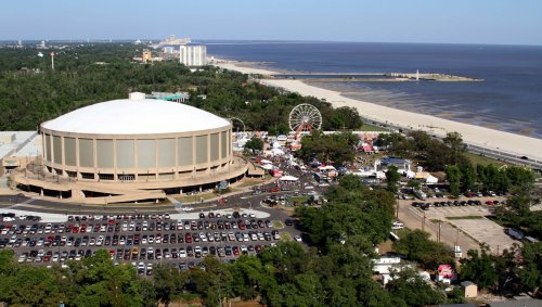 Aerial view of a large circular building near a beach, with a fairground and parking lot visible below.