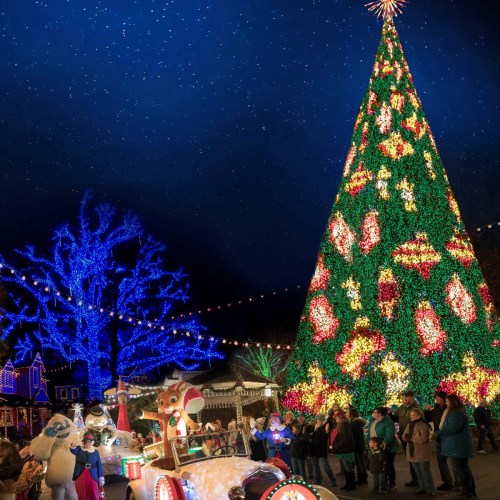 A festive scene with a large illuminated Christmas tree, colorful lights, and people enjoying holiday decorations.