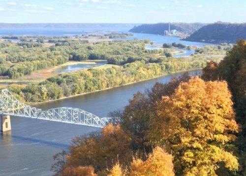 A scenic view of a river winding through autumn-colored trees and hills, with a bridge in the foreground.