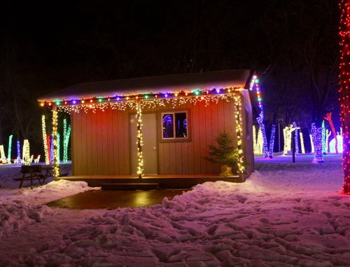 A cozy cabin adorned with colorful holiday lights, surrounded by snow and illuminated trees at night.