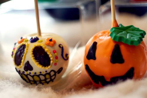 Two decorated cake pops: one resembling a sugar skull and the other a smiling pumpkin, set against a soft, fluffy background.