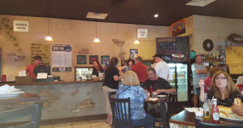 A busy restaurant interior with customers ordering at the counter and others dining at tables. Brightly decorated walls.