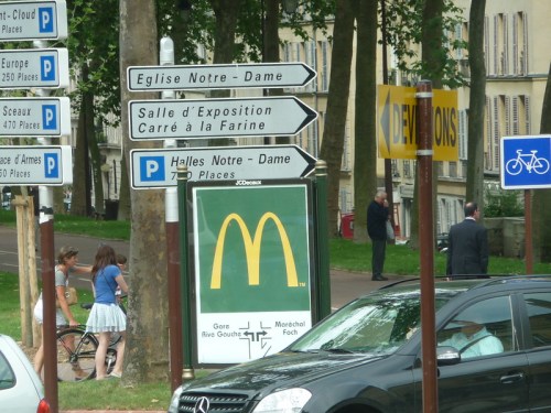 A street scene with directional signs, a McDonald's sign, and people walking and biking in a park-like area.