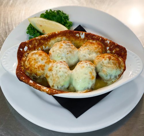 A dish of six cheesy baked dumplings in a crispy bowl, garnished with a lemon wedge and a side of greens.